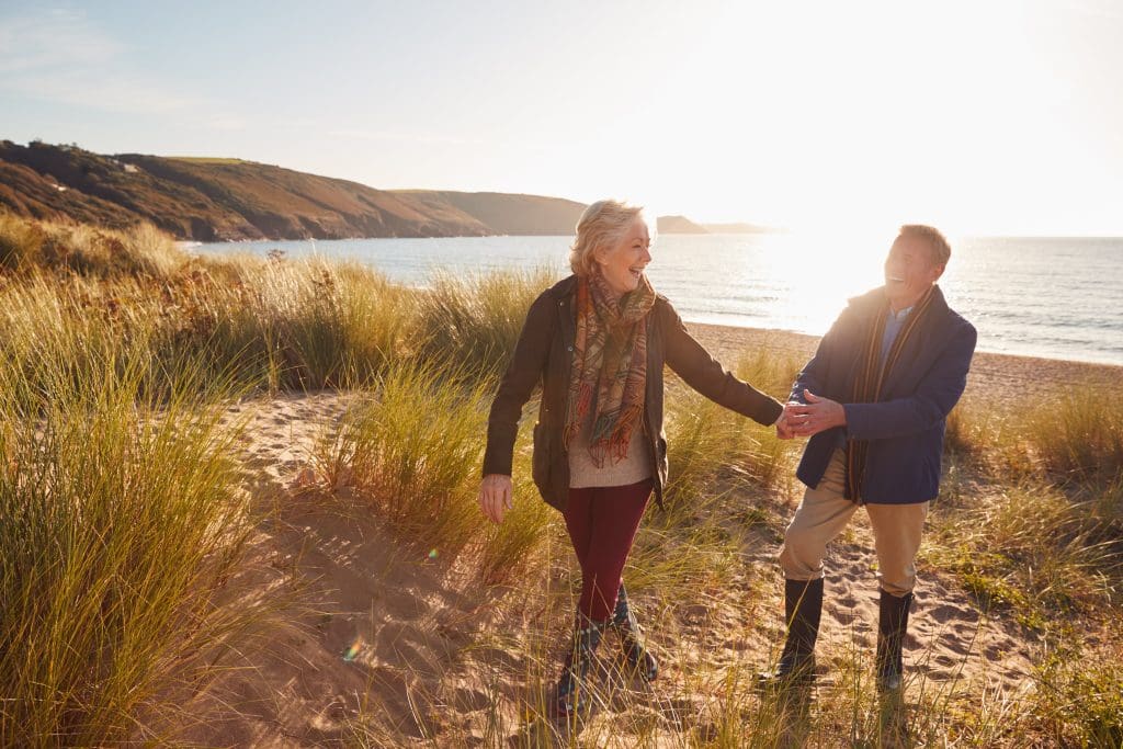 Older couple walking hand in hand on a beach at sunset, enjoying retirement.