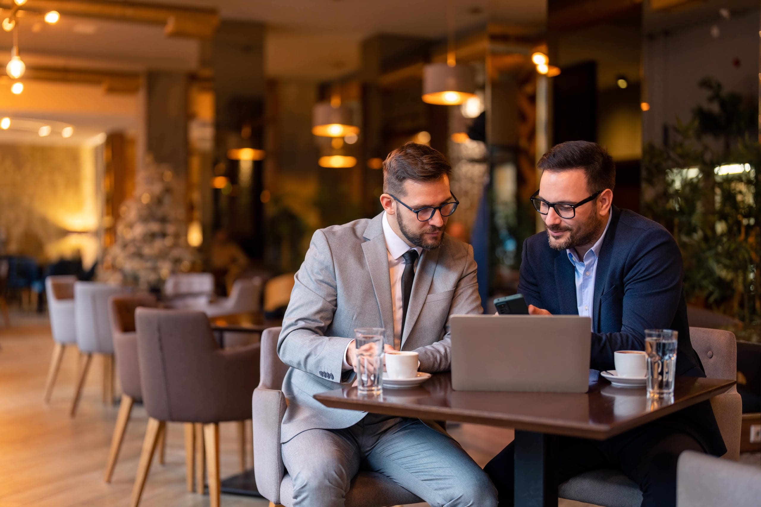 Two professionals in business attire are seated at a table in a café, collaborating over a laptop. One man is looking at a phone, while the other is writing in a notebook, indicating a business discussion.